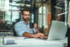 Morning Coffee. Portrait Of Young And Successful Bearded Man In Eyeglasses Holding Cup Of Coffee And Working With Laptop While Sitting At His Working Place